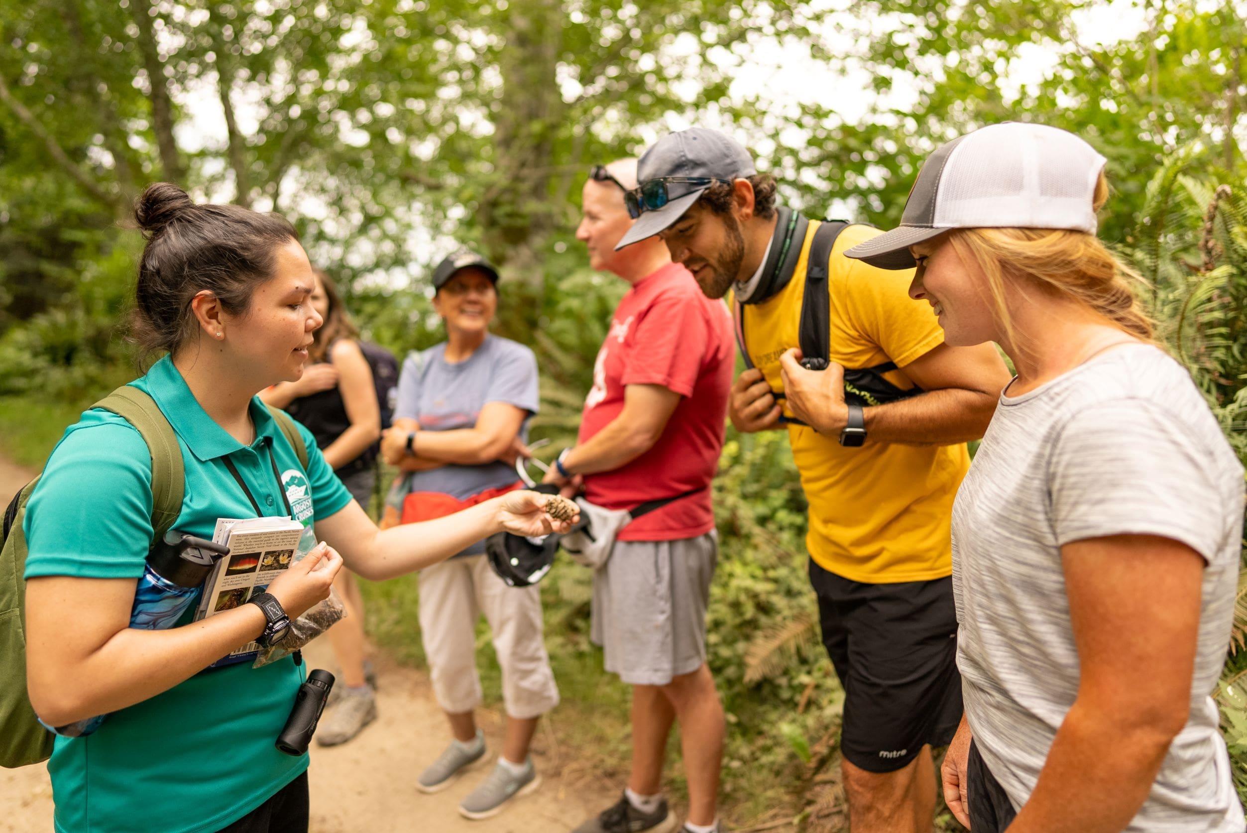 Woman holding pine cone with man & woman leaning forward to look at pinecone and people standing in background